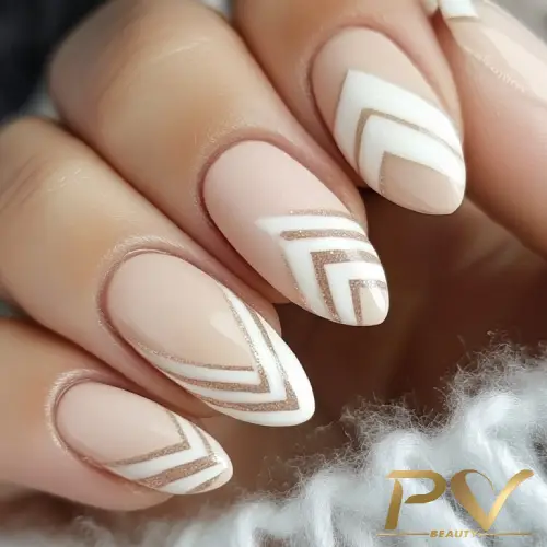 Unhas decoradas com esmalte nude, branco e dourado, apresentando padrões geométricos em forma de setas.