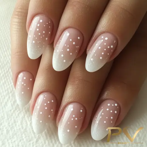 Mãos com unhas decoradas em estilo ombré branco e pontos brancos, sobre superfície clara.