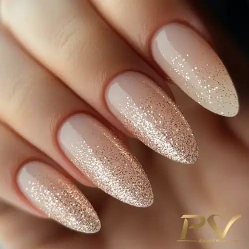 Unhas longas com esmalte nude e glitter dourado nas pontas, em um fundo neutro.