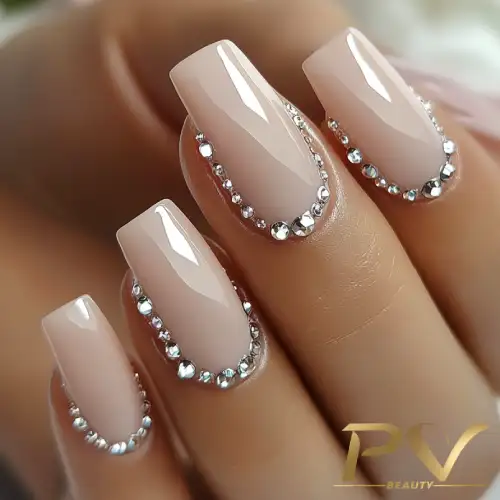 Unhas decoradas com esmalte nude e pequenas pedras brilhantes ao redor das bordas.