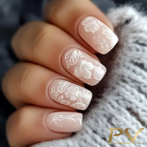 Unhas com esmalte nude e desenhos florais brancos, segurando tecido cinza texturizado.