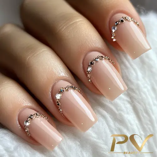 Unhas com esmalte nude e decoração de strass, formando arcos brilhantes sobre uma superfície branca.