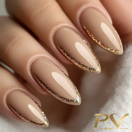 Unhas em formato stiletto com esmalte nude e bordas douradas com glitter.