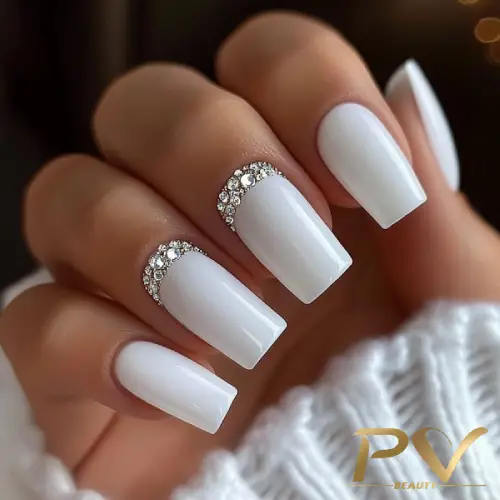 unhas-decoradas-branco-designer-de-unhas-pv-beauty-01 Mão com unhas brancas decoradas com pedras brilhantes, vestindo suéter branco.