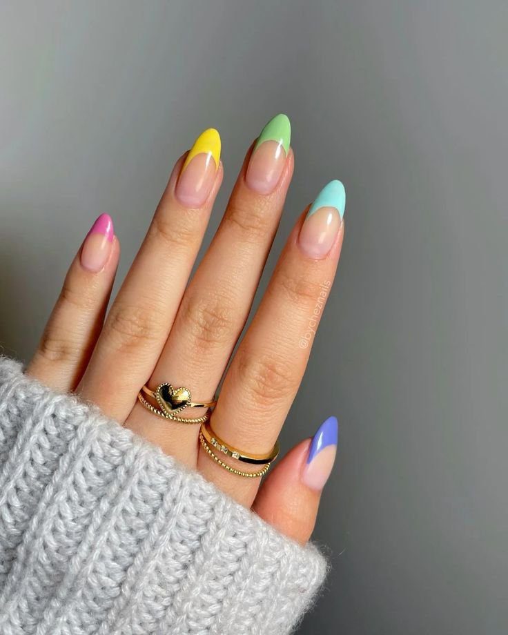 Mão com unhas coloridas em tons pastéis, usando anéis dourados e suéter cinza.