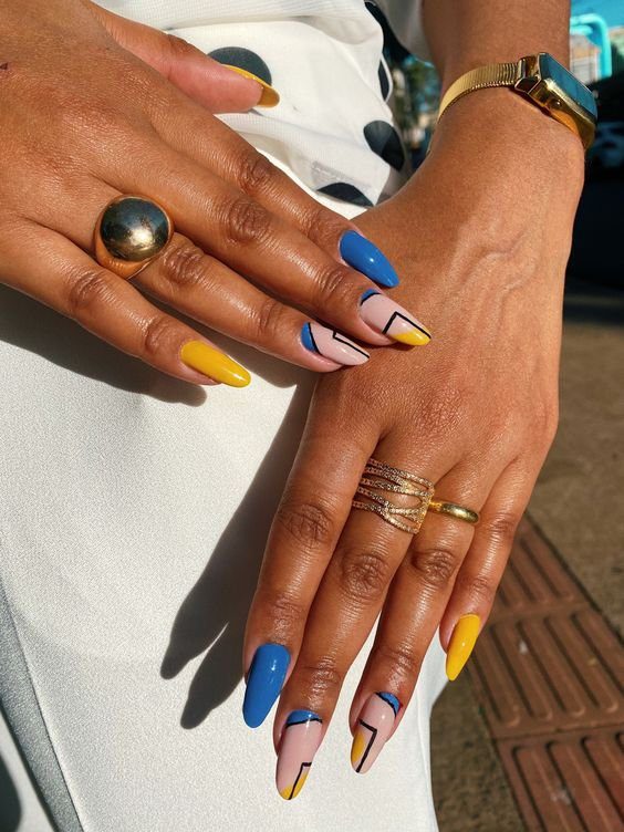unhas-decoradas-geometricas-pinterest Mãos com unhas decoradas em amarelo, azul e rosa, usando anéis e relógio dourado, sobre tecido branco.