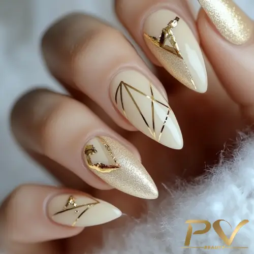 Unhas decoradas com esmalte nude e detalhes dourados geométricos e metálicos.