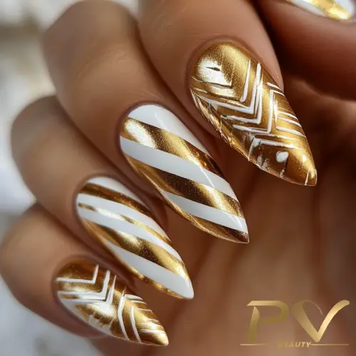 Unhas decoradas com esmalte dourado e branco, exibindo padrões geométricos e listras diagonais.