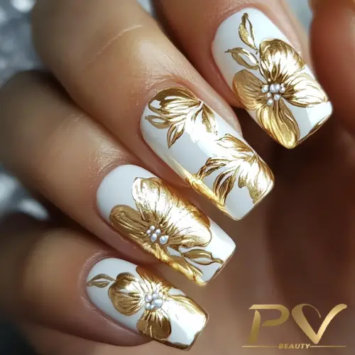 Unhas decoradas com esmalte branco e desenhos florais dourados, detalhes em pérolas.