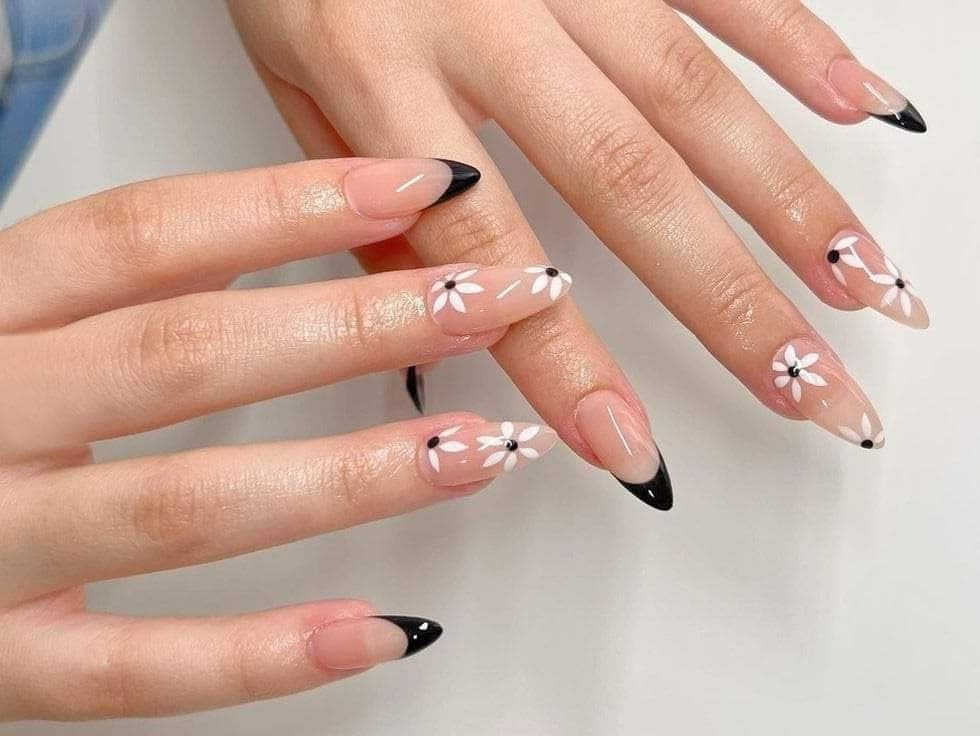 Mãos com unhas decoradas com pontas pretas e desenhos de flores brancas sobre base rosa clara.