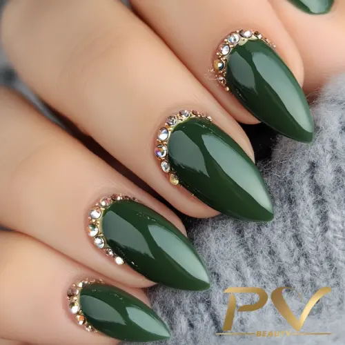 unhas-em-verde-strass-decoradas-pv-beauty-33 Unhas verdes brilhantes com pedrarias prateadas na base, sobre tecido cinza.