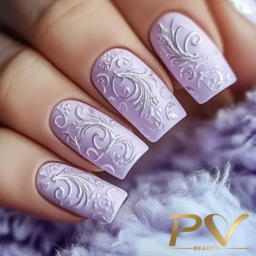 Unhas decoradas com esmalte lilás e design em relevo floral, sobre fundo desfocado em tons de roxo.