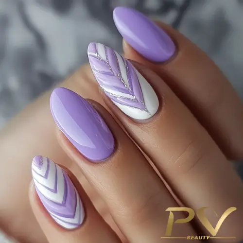 Unhas com esmalte lilás e branco, decoradas com desenhos geométricos e detalhes em brilho.