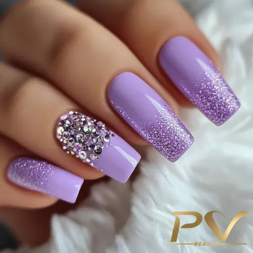 Unhas decoradas em tons de lilás, com glitter degradê e pedrarias brilhantes em uma das unhas.