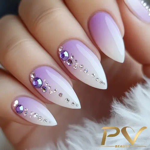 Unhas com esmalte degradê lilás para branco, decoradas com pedrarias brilhantes.