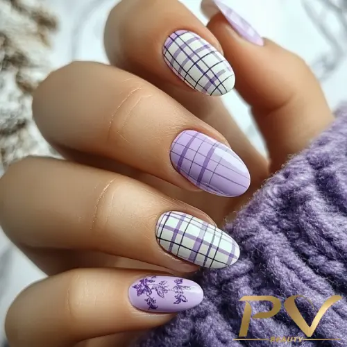 Mão com unhas decoradas em tons de roxo e branco, com padrões xadrez e florais, sobre tecido lilás.