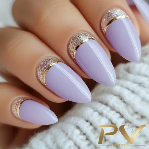 Unhas com esmalte lilás, detalhes dourados e glitter, sobre fundo de tecido claro.