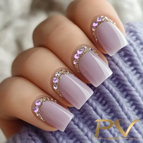 Unhas com esmalte lilás e detalhes de pedrarias douradas e roxas, sobre tecido lilás tricotado.