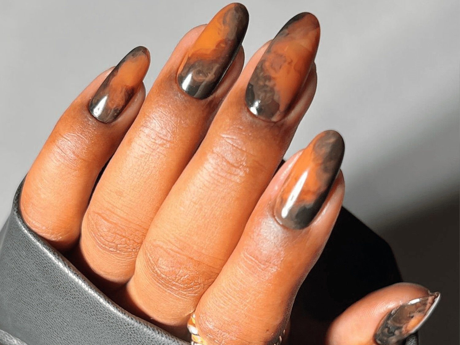 Mão com unhas longas e decoradas em padrão marmorizado nas cores laranja e preto, segurando tecido cinza.