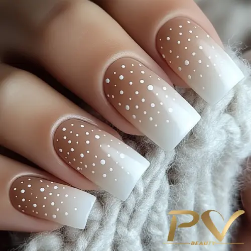 unhas-marrons-decoradas-poa-pv-beauty-16 Unhas decoradas com esmalte nude e pontinhos brancos, em fundo desfocado, segurando tecido cinza claro.