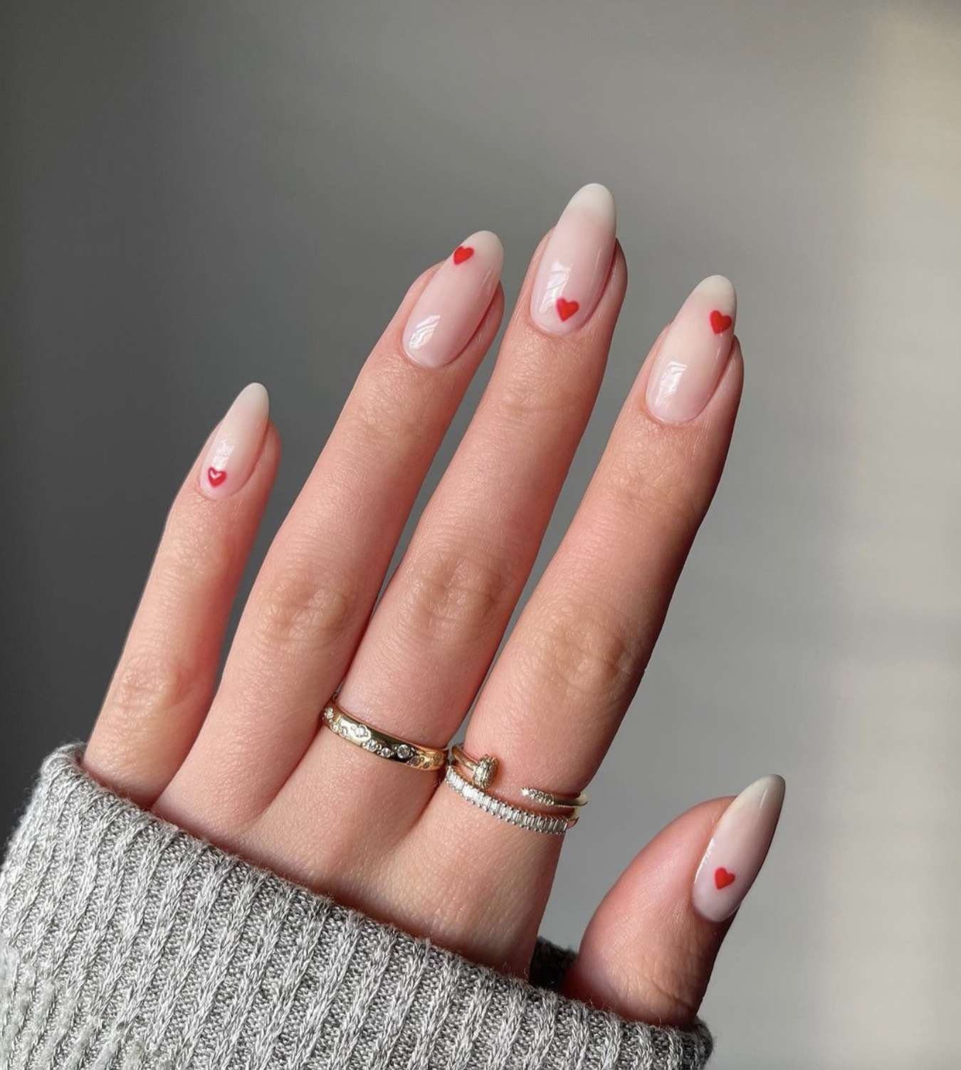 Mão com unhas decoradas com corações vermelhos e três anéis dourados, usando um suéter cinza.