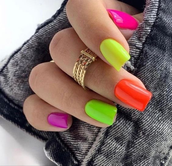 unhas-neon-sao-classicas-da-festividade Mão com unhas pintadas em cores neon: rosa, verde, amarelo e laranja, segurando tecido jeans e usando anel dourado.
