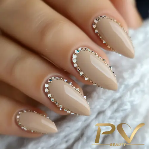 unhas-nude-strass-glamour-decoradas-esmaltacao-pv-beauty-33 Unhas com esmalte nude decoradas com pequenas pedras brilhantes ao redor das bordas, sobre superfície clara.