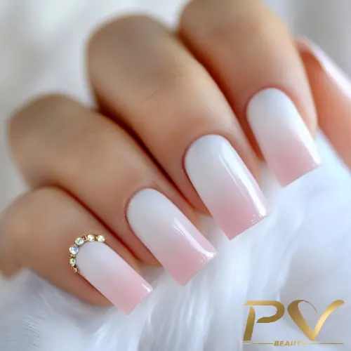 Unhas compridas com esmalte em degradê branco e rosa, detalhe de anel dourado com pedras brilhantes em um dedo.