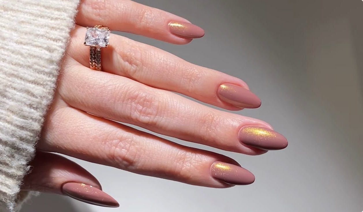 Mão com unhas pintadas de rosa com brilho dourado e anel com pedra grande em destaque.