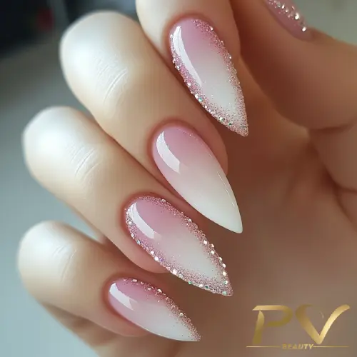 Unhas longas e pontudas com esmalte em degradê rosa e branco, decoradas com glitter prateado nas bordas.