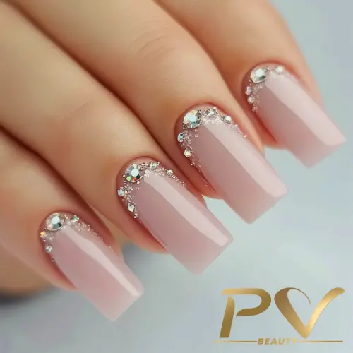 Unhas quadradas com esmalte rosa claro e decoração de strass prateados nas cutículas.