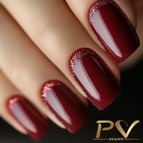 unhas-vinho-glitter-elegante-decoradas-pv-beauty-33 Unhas com esmalte vermelho escuro e detalhes de glitter próximo às cutículas, fundo desfocado.