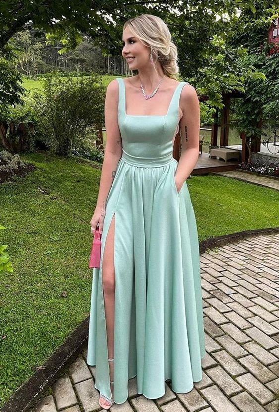 Mulher em vestido verde claro com fenda, segurando uma bolsa rosa, em ambiente externo com &aacute;rvores e gramado.