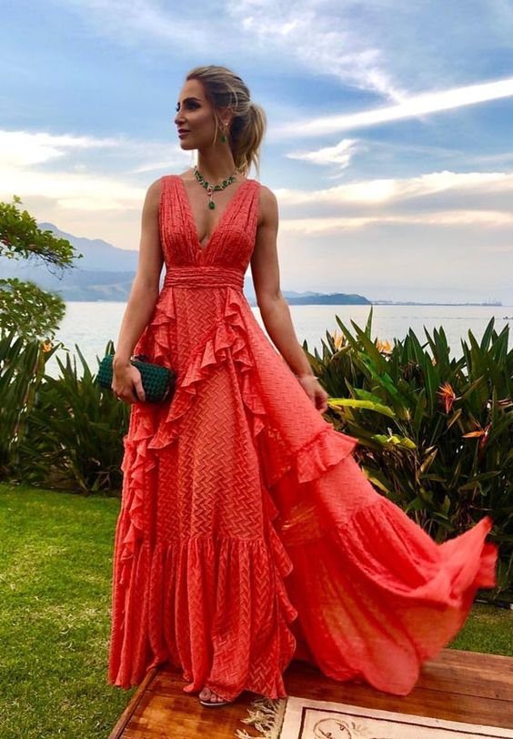 Pessoa em vestido coral longo em jardim &agrave; beira-mar, com c&eacute;u parcialmente nublado ao fundo.