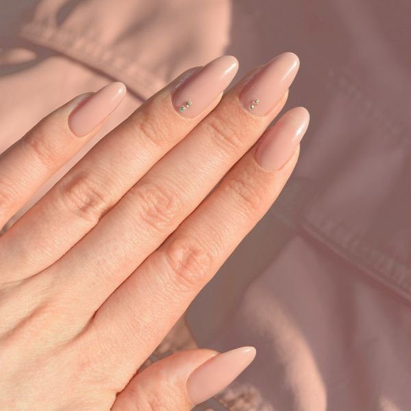 unha-clean-amendoada-brilhos Mão com unha amendoada clean em tom nude e brilhos esverdeados no dedo anelar e no dedo do meio.